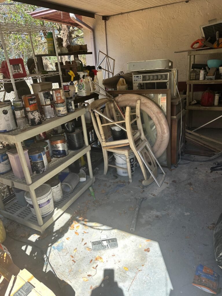 Covered outdoor storage area with paint cans, chairs, and AC units before cleanout