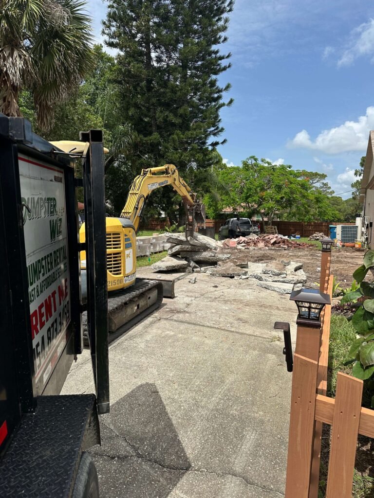 Concrete driveway mid-demolition Palm Bay FL — excavator breaking slabs with crew member supervising
